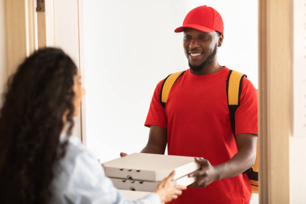 Fast Food Delivery Concept. Cheerful African American male courier wearing yellow thermo backpack and red uniform holding pizza boxes from restaurant and giving it to female customer standing at door